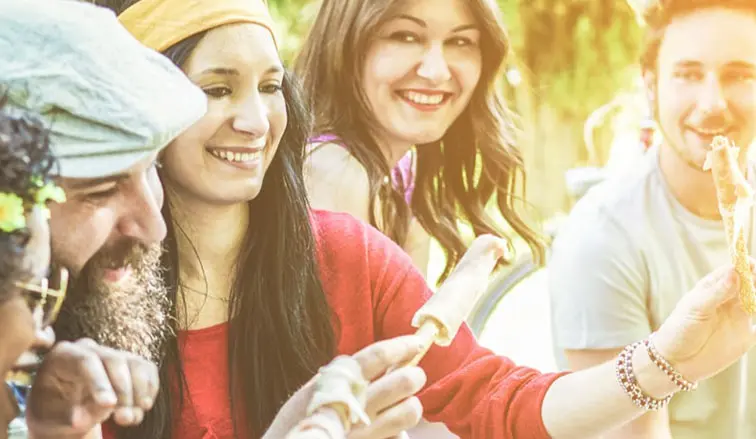 A group of friends enjoy a lively outdoor gathering, sharing food and laughter under warm sunlight, with greenery in the background.