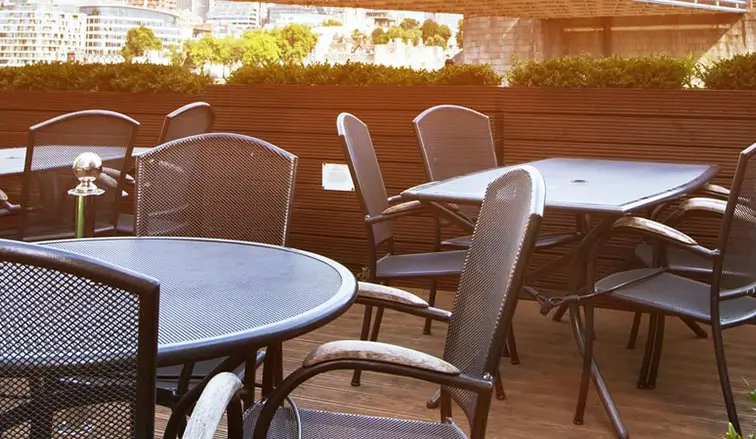 Outdoor patio with black metal chairs and tables, surrounded by green hedges and a view of a cityscape in the background.