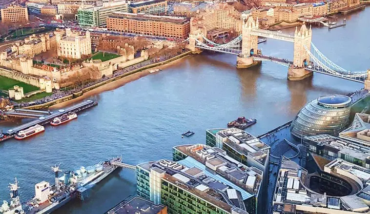 Aerial view of London's Tower Bridge over the Thames River, with historic and modern buildings surrounding the waterway.