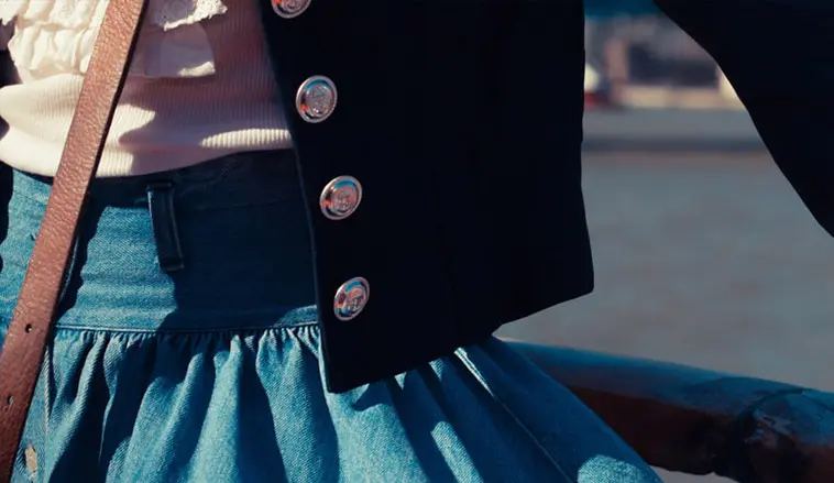 A close-up of a stylish outfit featuring a black jacket with shiny buttons and a blue denim skirt, with a blurred background.