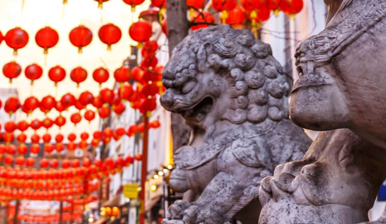 Two stone lion statues guard a vibrant street adorned with red lanterns, creating a festive and cultural atmosphere.