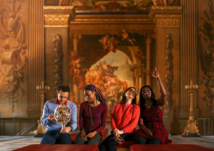A group of four people poses in an opulent interior, featuring a detailed mural and ornate decorations, with one person holding a globe.