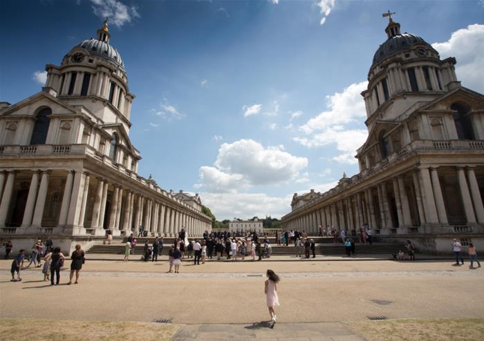 A girl in a pink dress walks towards historic buildings under a blue sky, with crowds gathered between ornate columns.