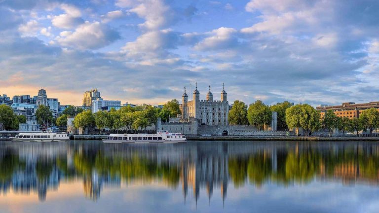 Thames River Sightseeing Blog 5 The Tower of London stands majestically by the river, mirrored in calm waters, framed by lush trees and a vibrant city skyline.