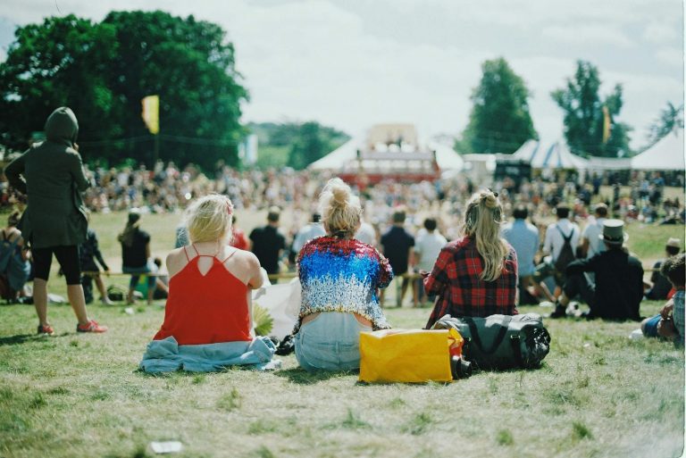 Three women in trendy outfits sit on the grass, enjoying a lively festival crowd on a sunny day, with colorful tents in the background.