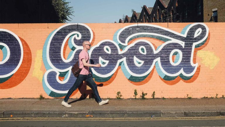 A person walks past a vibrant mural that spells "Good" in bold, colorful letters on a brick wall under a clear blue sky.
