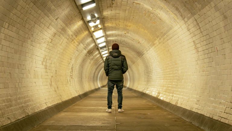 A person stands alone in a long, dimly-lit tunnel with white tiled walls, emanating a sense of isolation and contemplation.