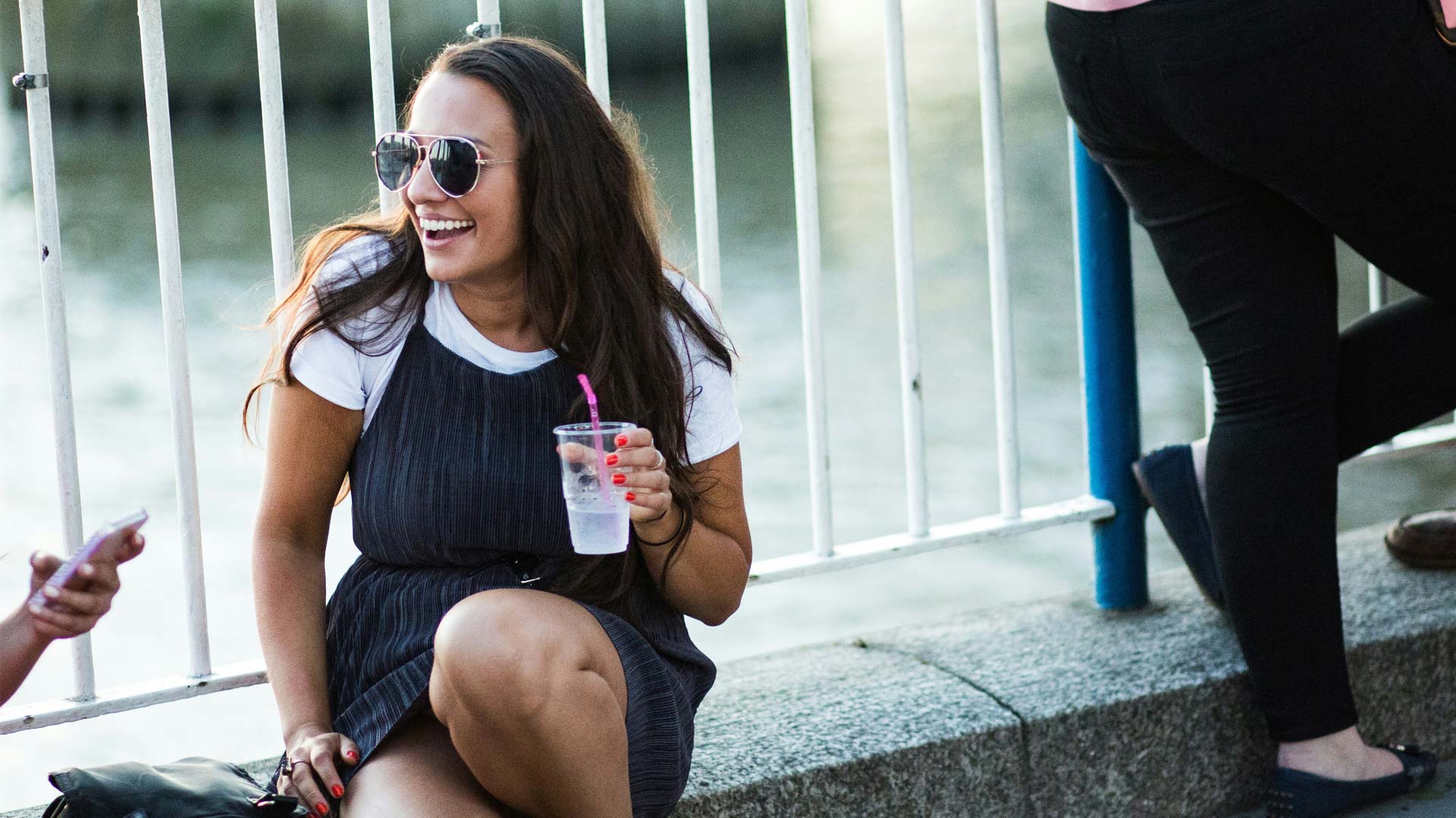 A young woman sits by a waterfront, holding a drink with a pink straw, dressed in a white t-shirt and a dark dress.