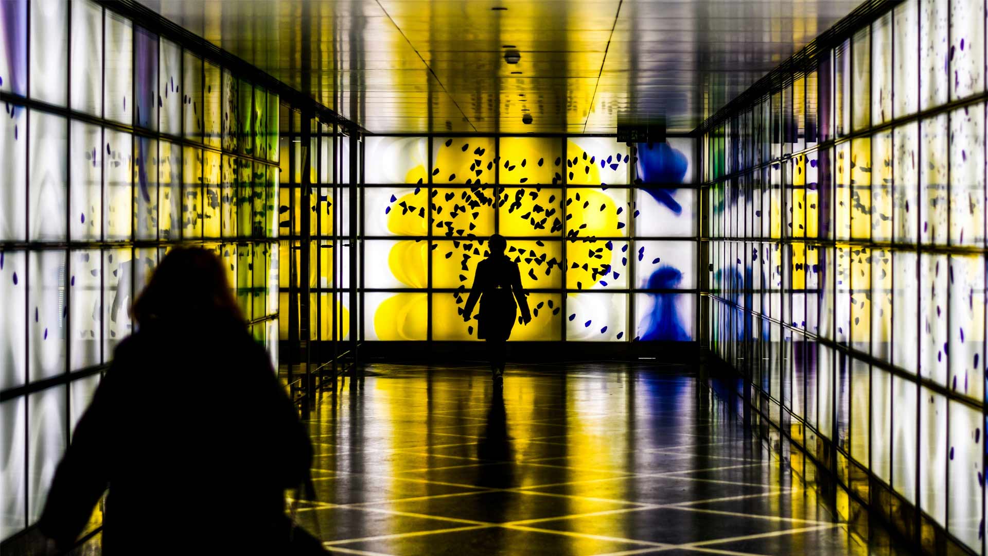 Silhouette of a person walking through a brightly lit corridor featuring colorful, abstract wall art in yellow, blue, and purple.