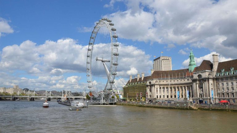 The London Eye towers over the Thames, with cloudy blue skies, charming boats, and historic buildings lining the riverside.