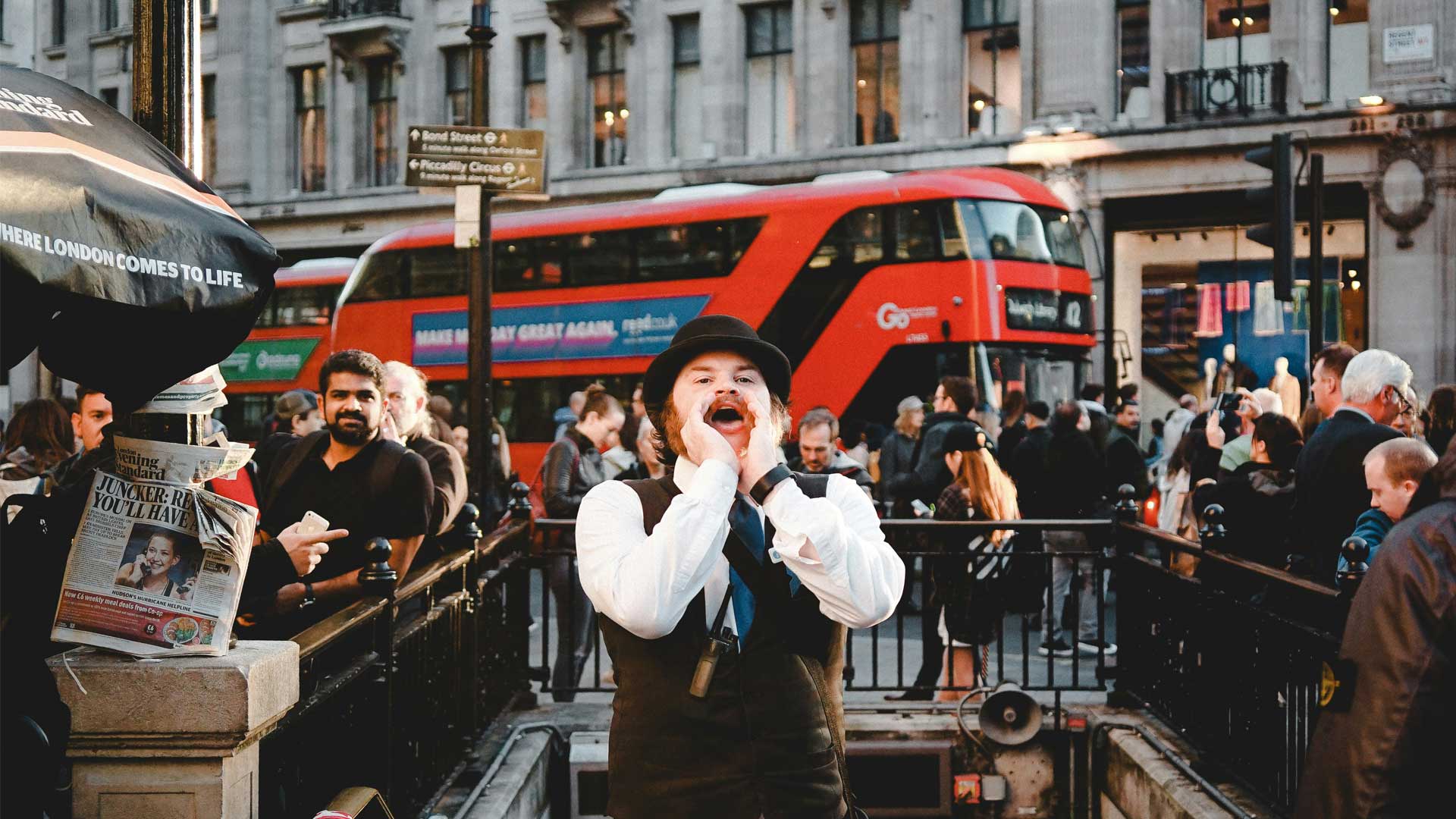 A bustling London street scene with a red double-decker bus in the background, surrounded by pedestrians and a newspaper stand.