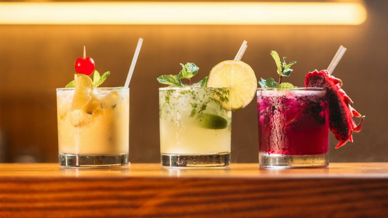 Three colorful cocktails on a wooden bar: a yellow drink with a cherry, a minty lime drink, and a vibrant pink drink with dragon fruit.