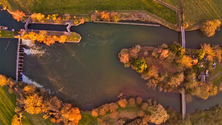 Aerial view of a winding river with autumn-colored trees, a small island, and a bridge connecting the banks, creating a tranquil landscape.
