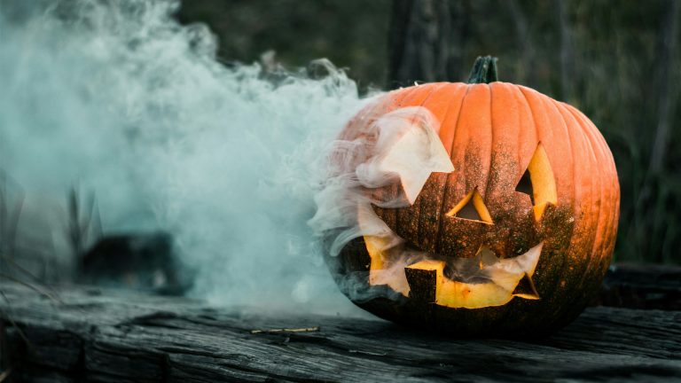 A carved Halloween pumpkin emits wisps of smoke, resting on a rustic wooden log against a blurred forest backdrop.