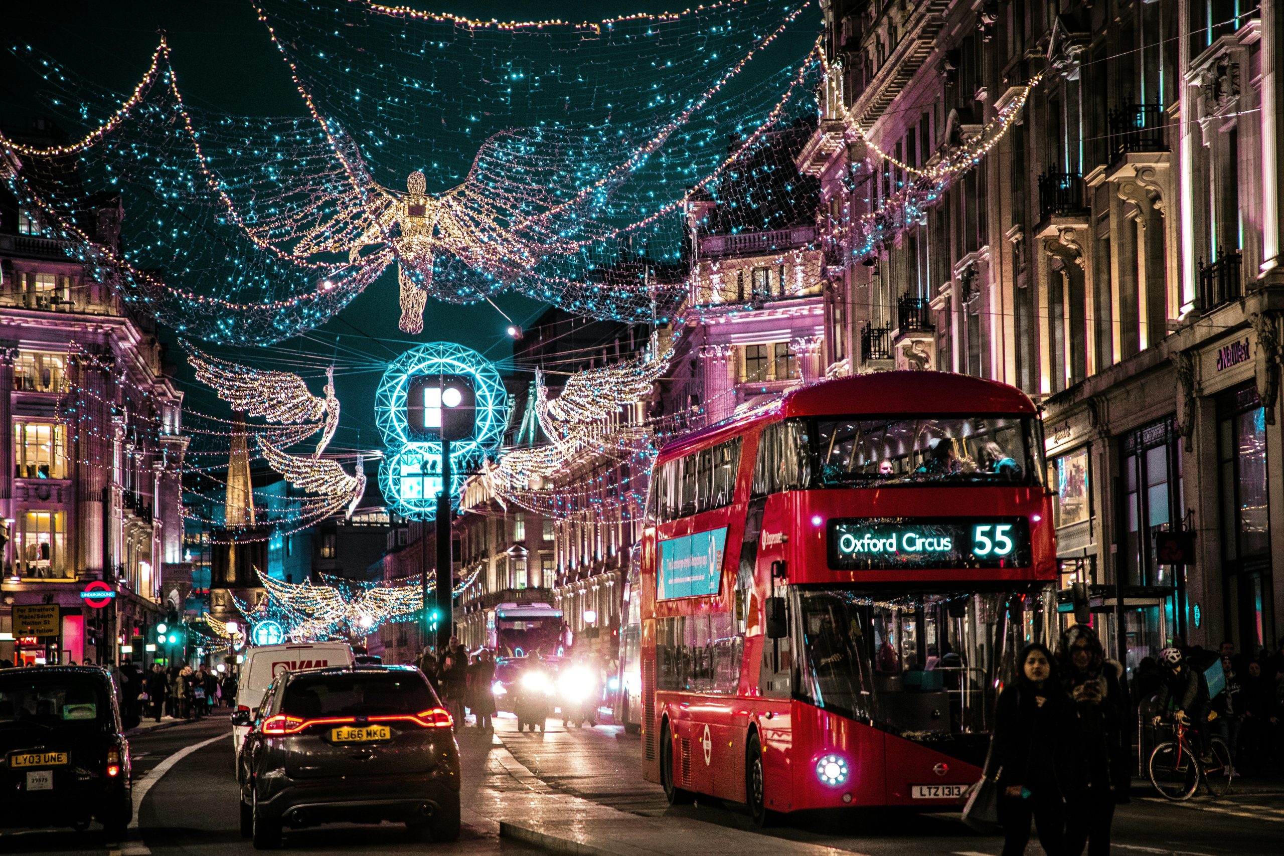 A busy street at night illuminated by festive lights, featuring a red double-decker bus labeled "Oxford Circus 55" and bustling traffic.