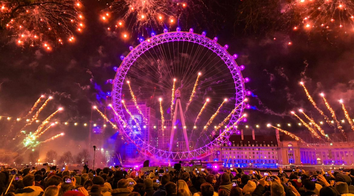 A vibrant fireworks display illuminates the London Eye and the night sky, surrounded by a crowd of spectators capturing the moment.