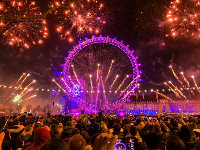 A vibrant fireworks display illuminates the London Eye and the night sky, surrounded by a crowd of spectators capturing the moment.
