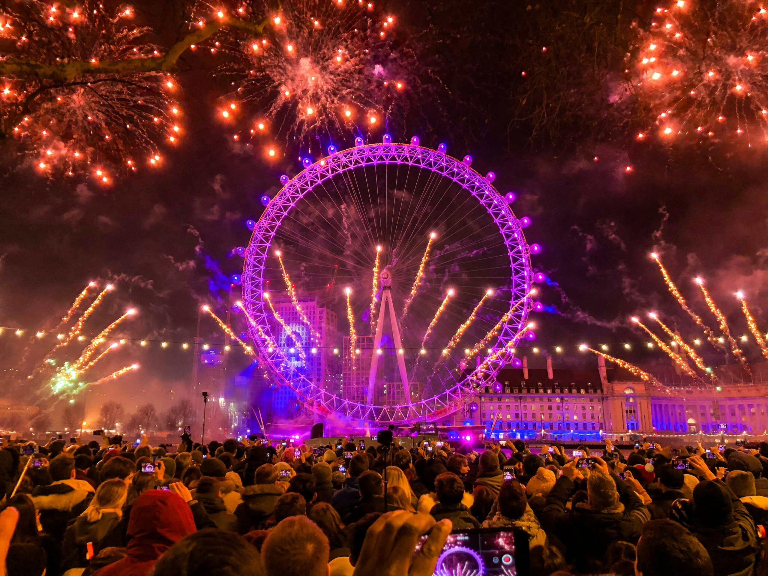 A vibrant fireworks display illuminates the London Eye and the night sky, surrounded by a crowd of spectators capturing the moment.