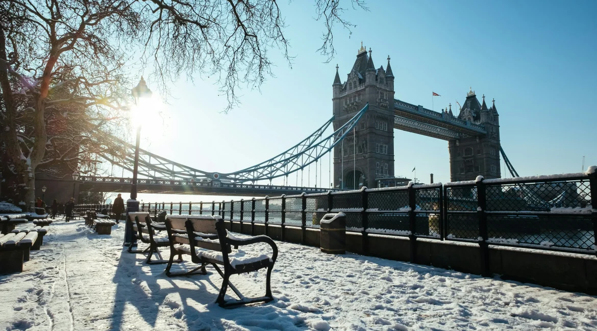 Snowy scene near Tower Bridge with benches, a glistening river, and softly glowing sunlight in a clear blue sky.