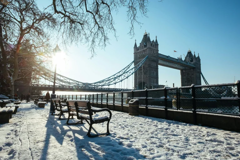 Snowy scene near Tower Bridge with benches, a glistening river, and softly glowing sunlight in a clear blue sky.