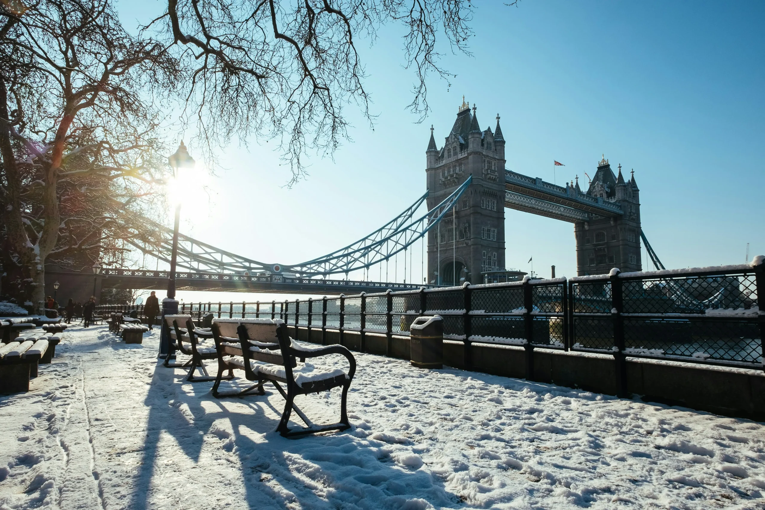 Snowy scene near Tower Bridge with benches, a glistening river, and softly glowing sunlight in a clear blue sky.