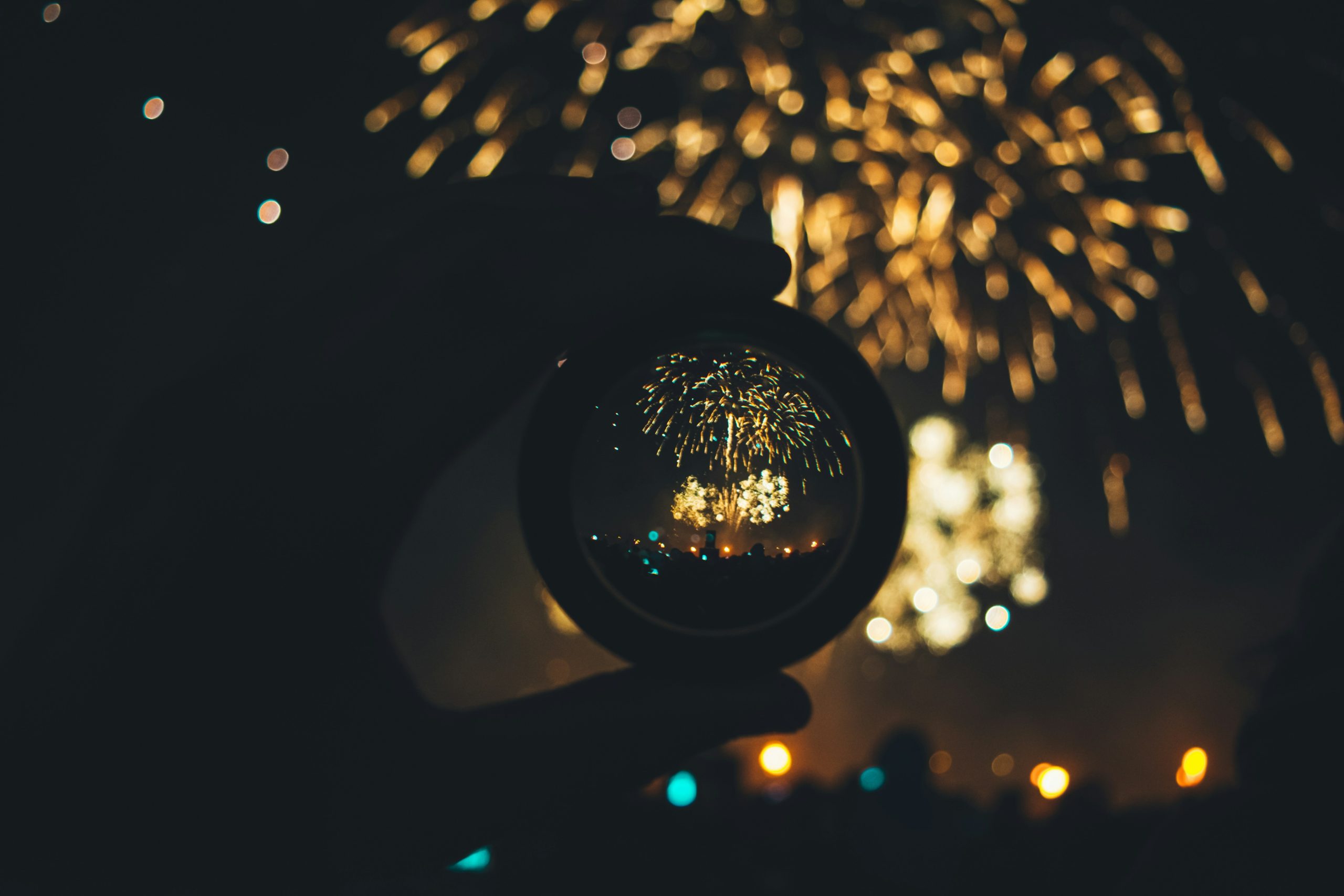 A hand holding a circular lens reflects vibrant fireworks bursting in the night sky, surrounded by a captivated crowd.