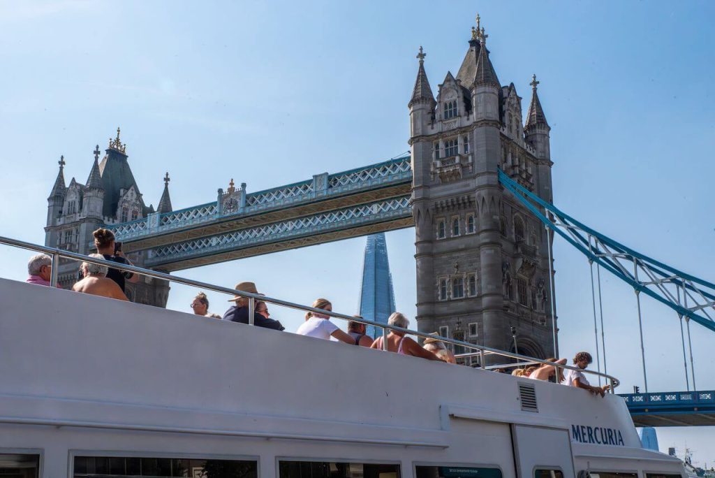 A crowded boat beneath Tower Bridge, with the Shard skyscraper visible in the background against a clear blue sky.