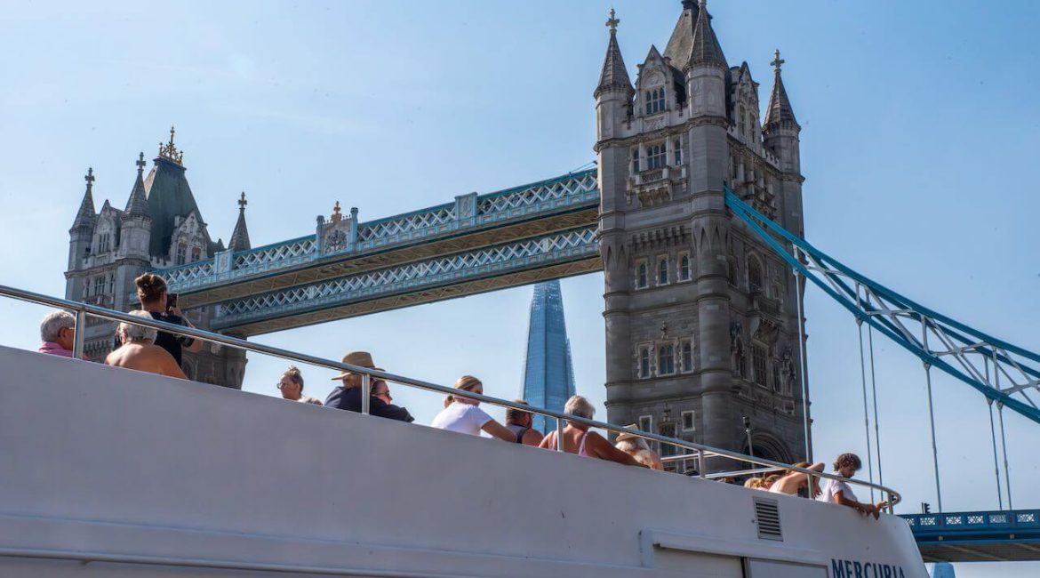 A crowded boat beneath Tower Bridge, with the Shard skyscraper visible in the background against a clear blue sky.