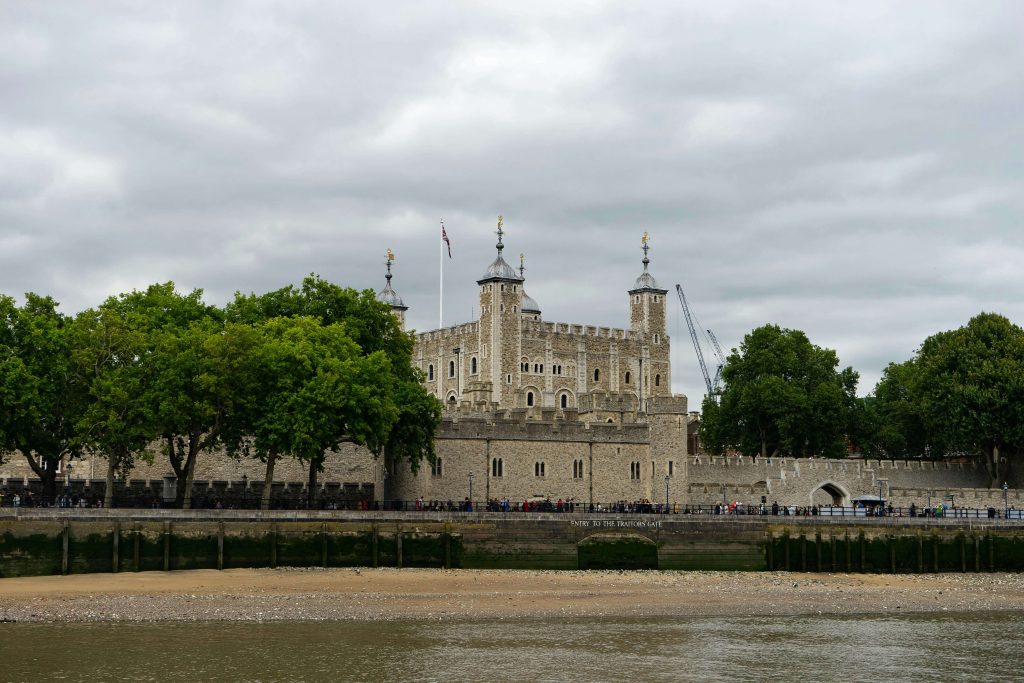 The Tower of London stands tall by the River Thames, surrounded by lush trees and cloudy skies, showcasing its historic architecture.