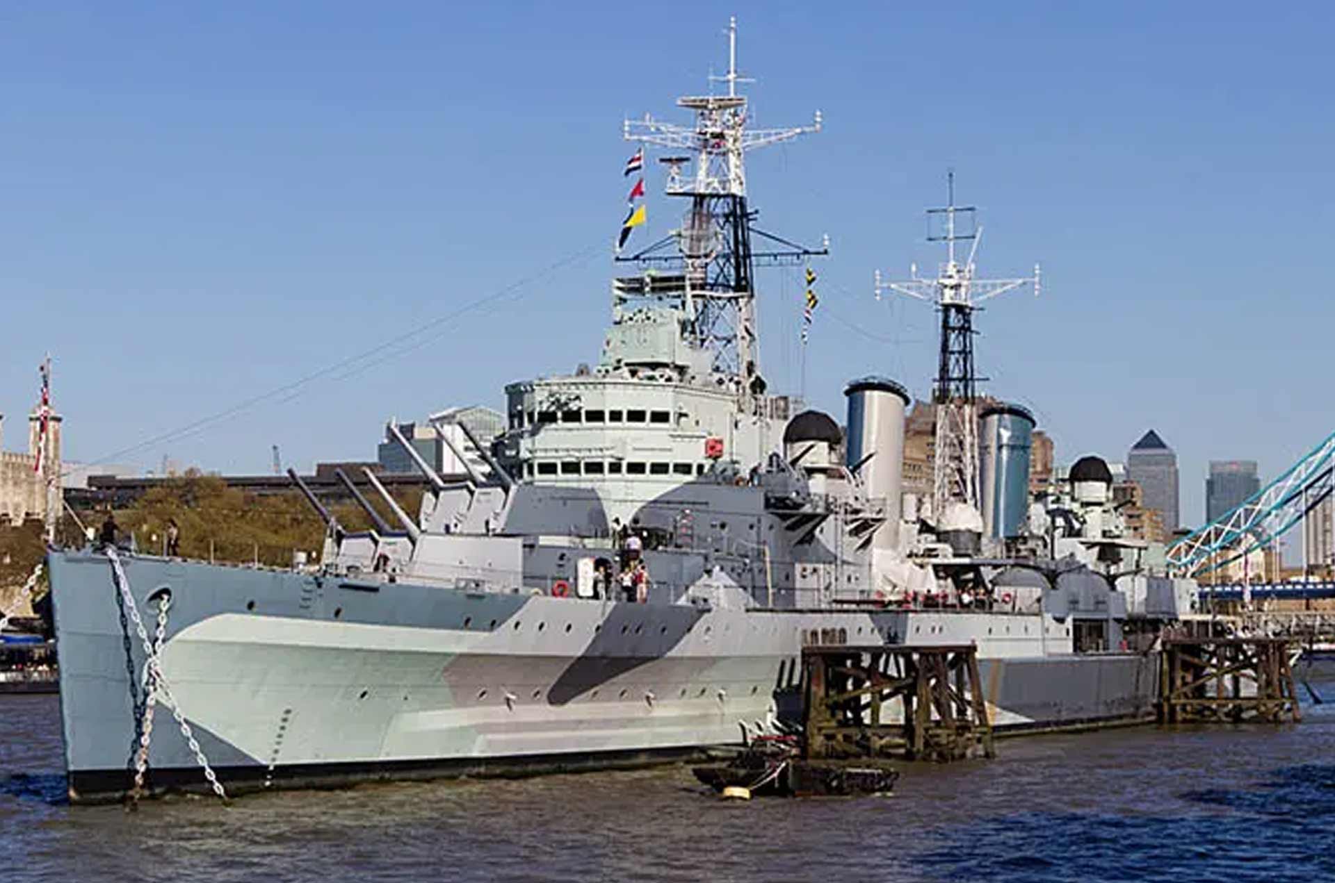 A historic warship docked in calm waters, showcasing gray camouflage and naval structures under a blue sky with scattered clouds.