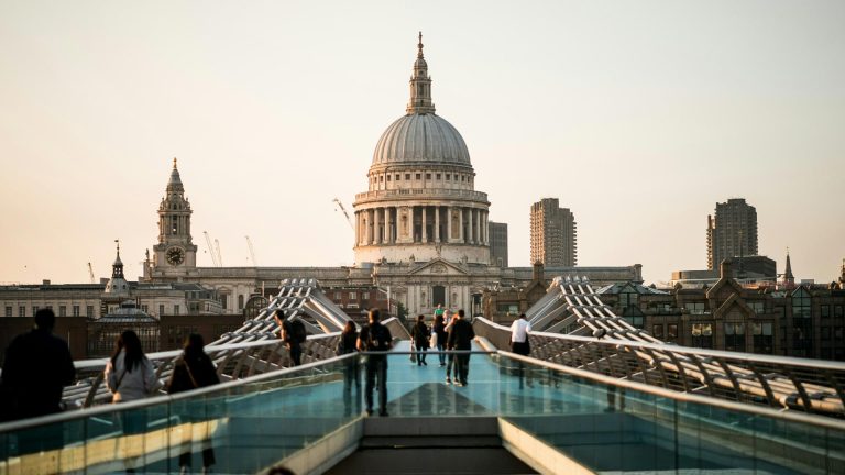 People stroll across a modern bridge, with St. Paul's Cathedral's dome and city skyline bathed in the warm glow of sunset.