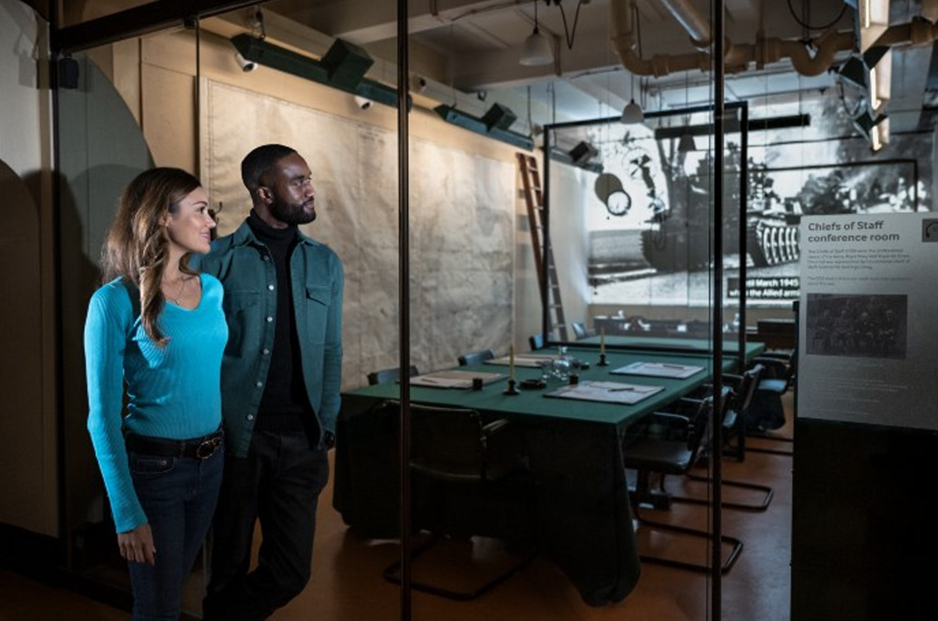 Visitors stand in a historic conference room, featuring a green table, chairs, and a projector displaying black-and-white military images.