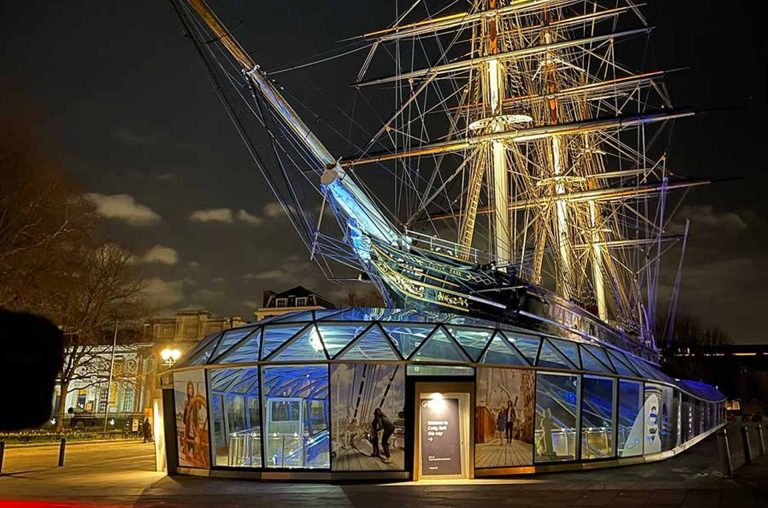 A historic ship stands illuminated at night, partially enclosed in a glass structure, showcasing its masts and intricate details.