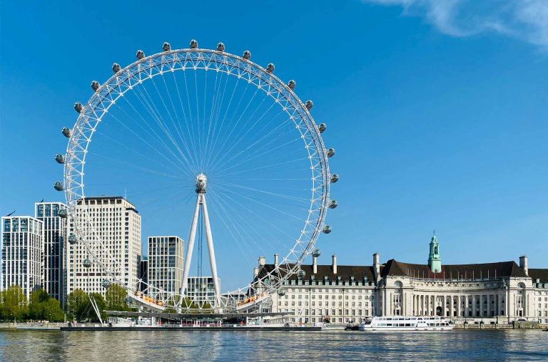 A view of the London Eye against a clear blue sky, beside modern buildings and the historic County Hall along the Thames River.