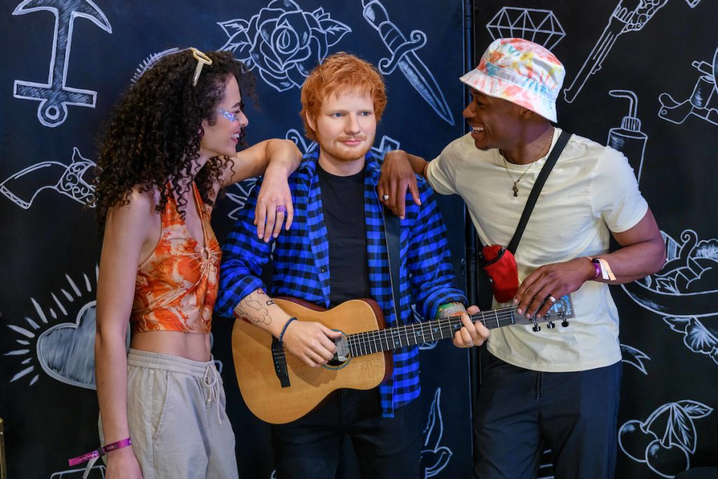 Ed Sheran holds an acoustic guitar, flanked by two friends, with a colorful graffiti-style backdrop filled with artistic designs.