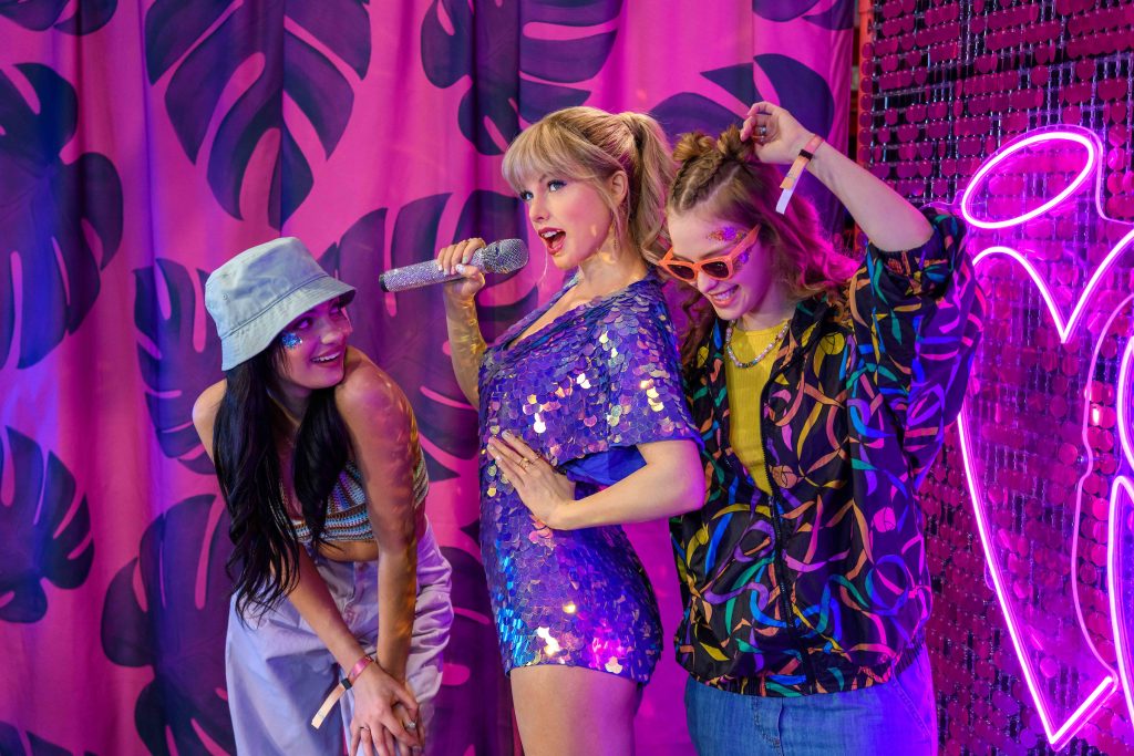 Three friends pose playfully with Tailor against a vibrant pink background with palm leaves, surrounded by colorful lights and a neon heart.