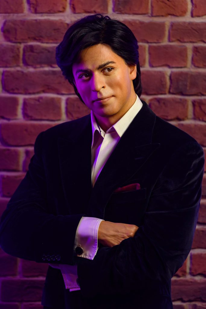 A person in a velvet blazer crossed their arms, standing against a brick wall illuminated with purple light.