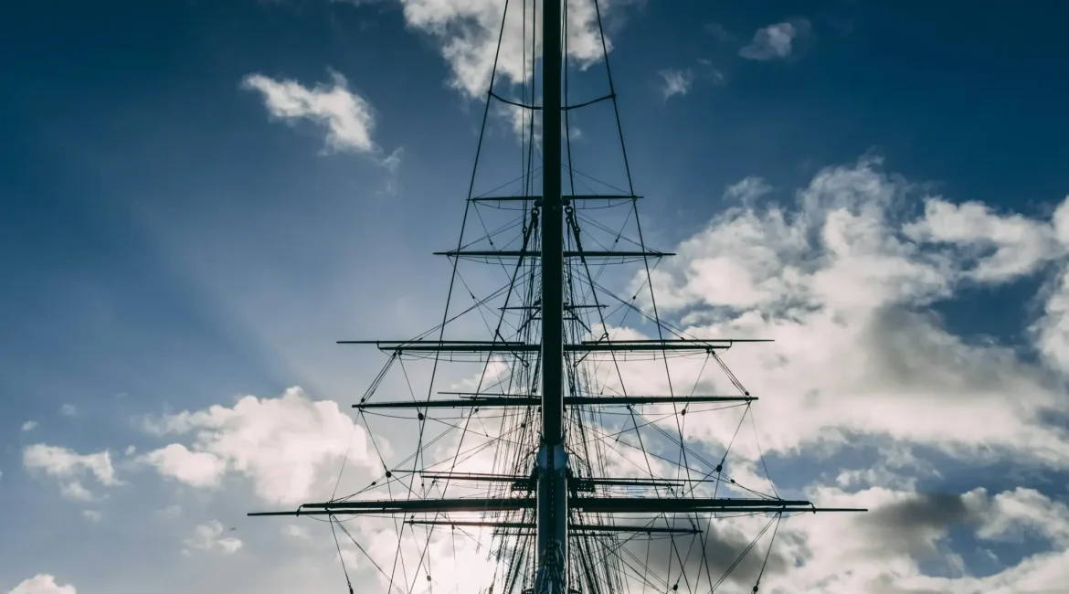 Silhouette of a tall ship's masts and rigging against a bright sky with wispy clouds, capturing a serene maritime atmosphere.