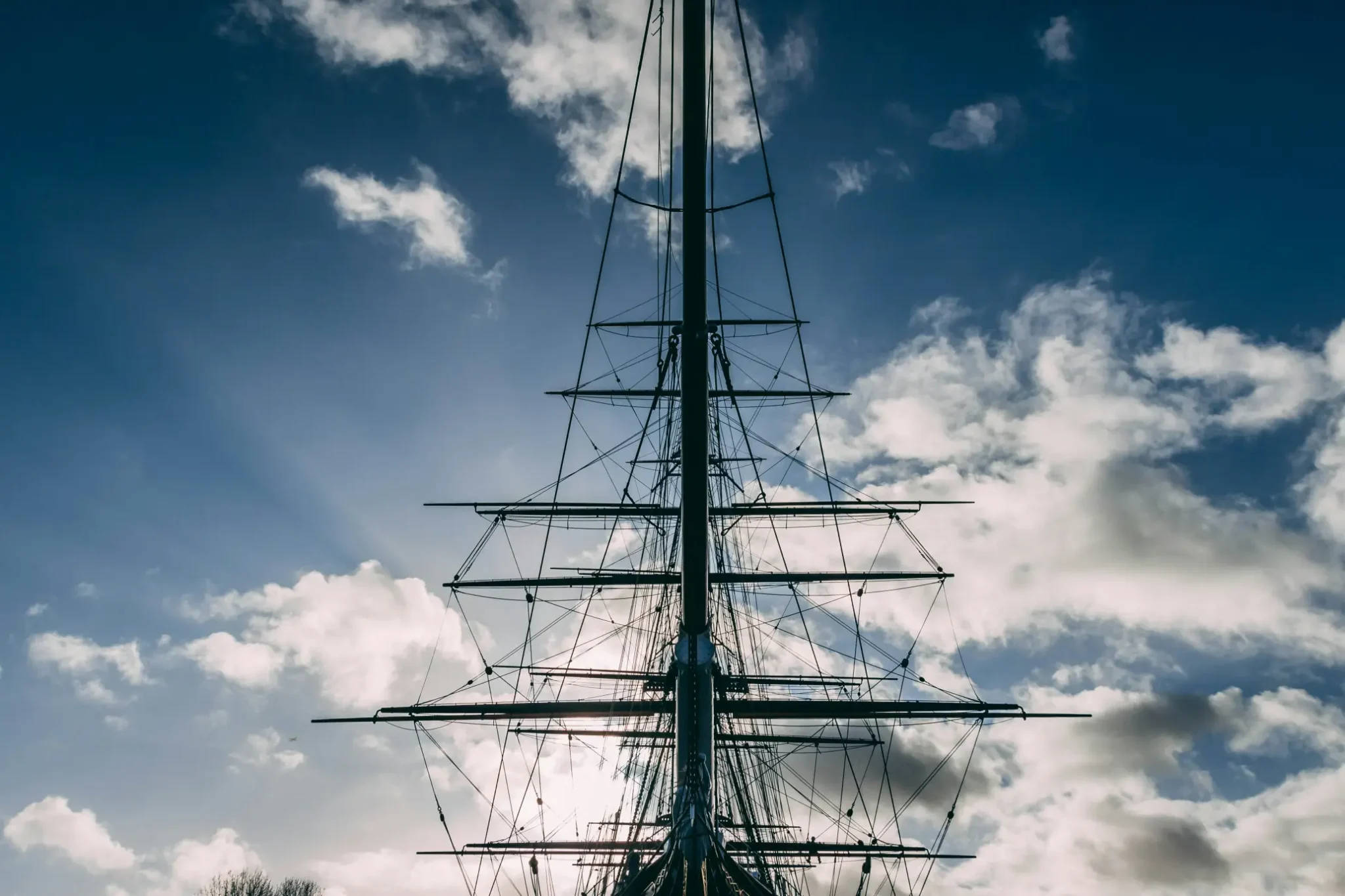 Silhouette of a tall ship's masts and rigging against a bright sky with wispy clouds, capturing a serene maritime atmosphere.