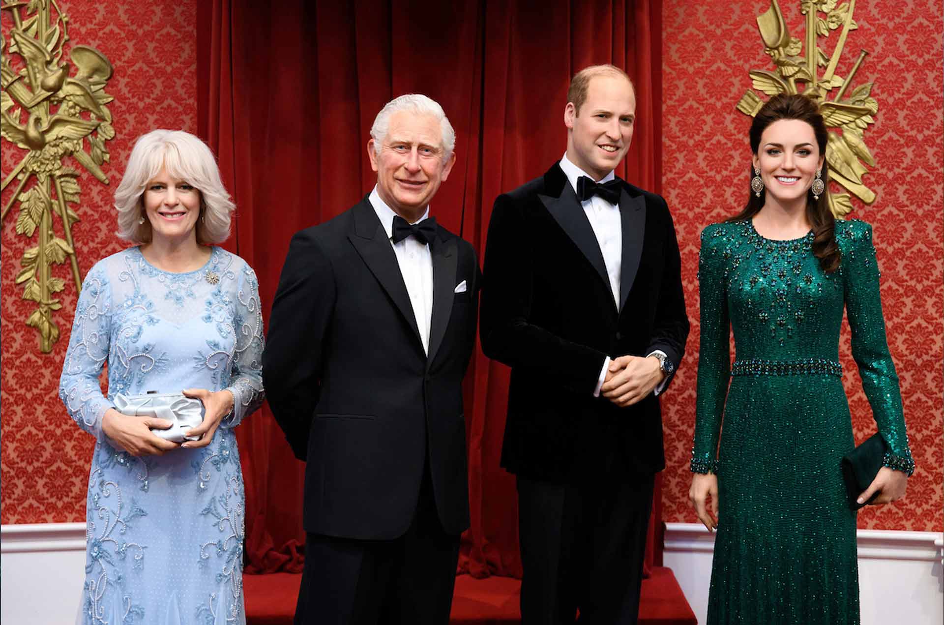 A formal gathering features four elegantly dressed individuals against a rich red backdrop with ornate gold decor.