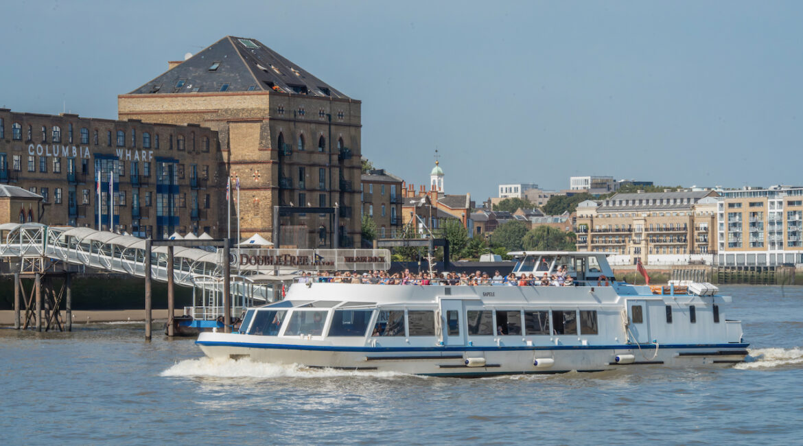 Sightseeing boat on a Thames River cruise passing historic buildings and piers on a sunny day in London.