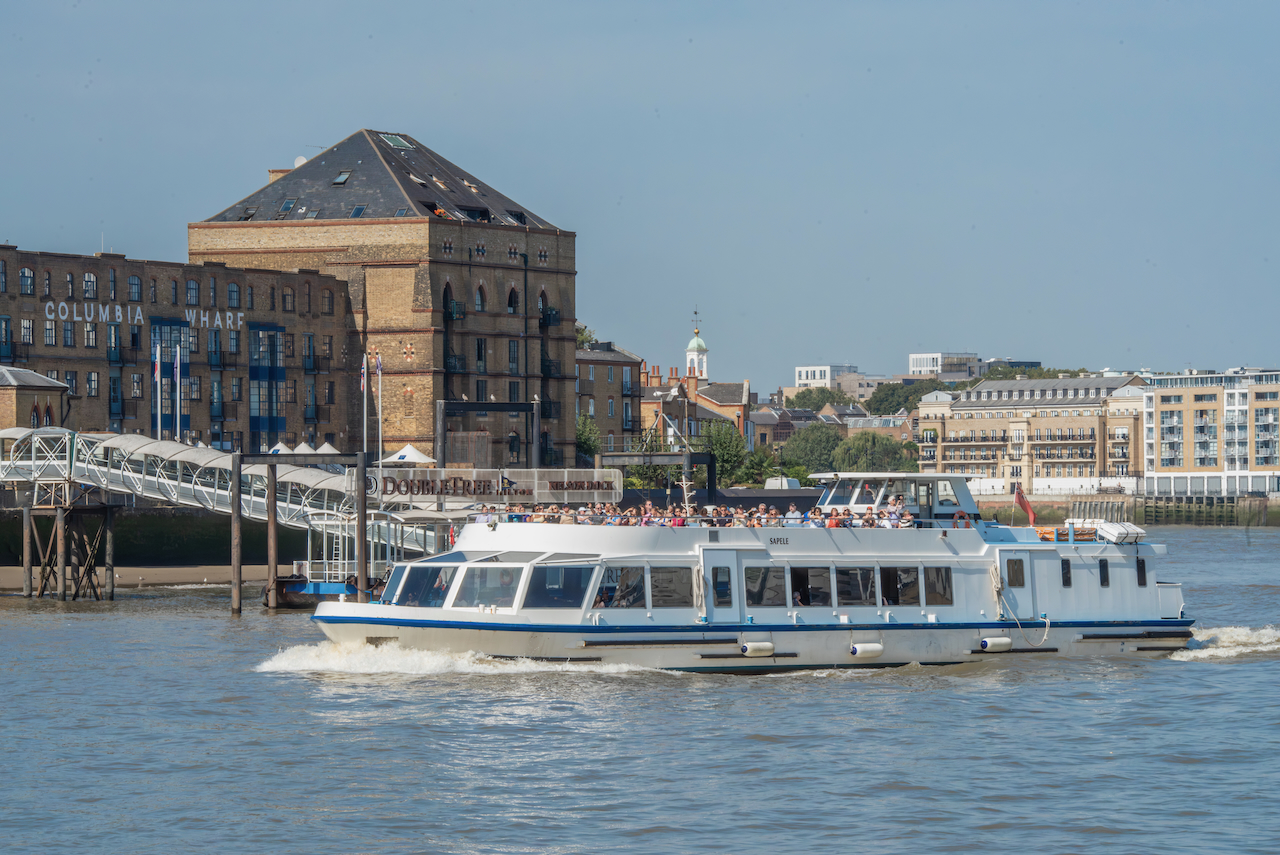 Sightseeing boat on a Thames River cruise passing historic buildings and piers on a sunny day in London.