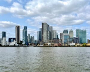 The modern skyscrapers of Canary Wharf skyline viewed from a boat on the River Thames under a cloudy blue sky.