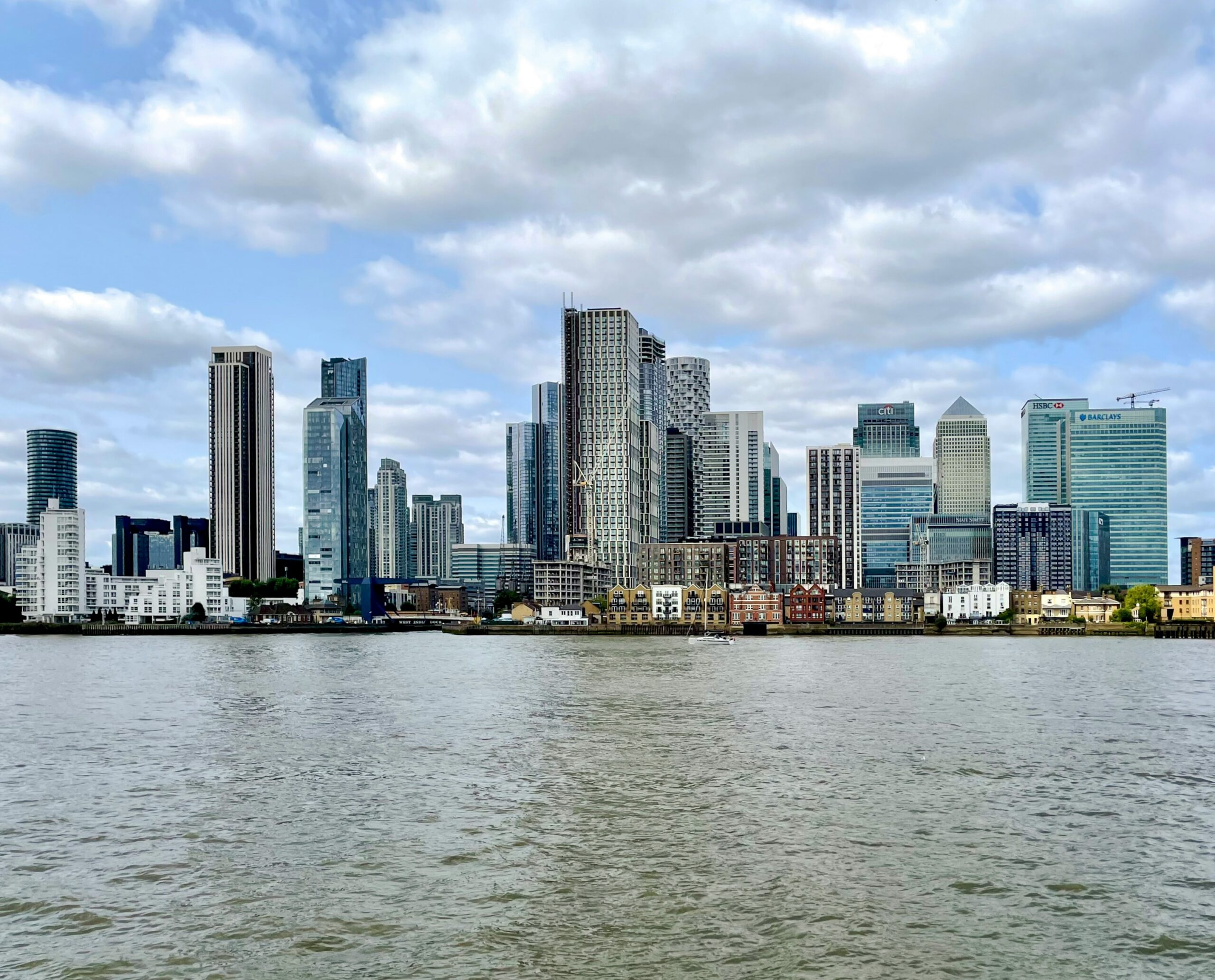 The modern skyscrapers of Canary Wharf skyline viewed from a boat on the River Thames under a cloudy blue sky.
